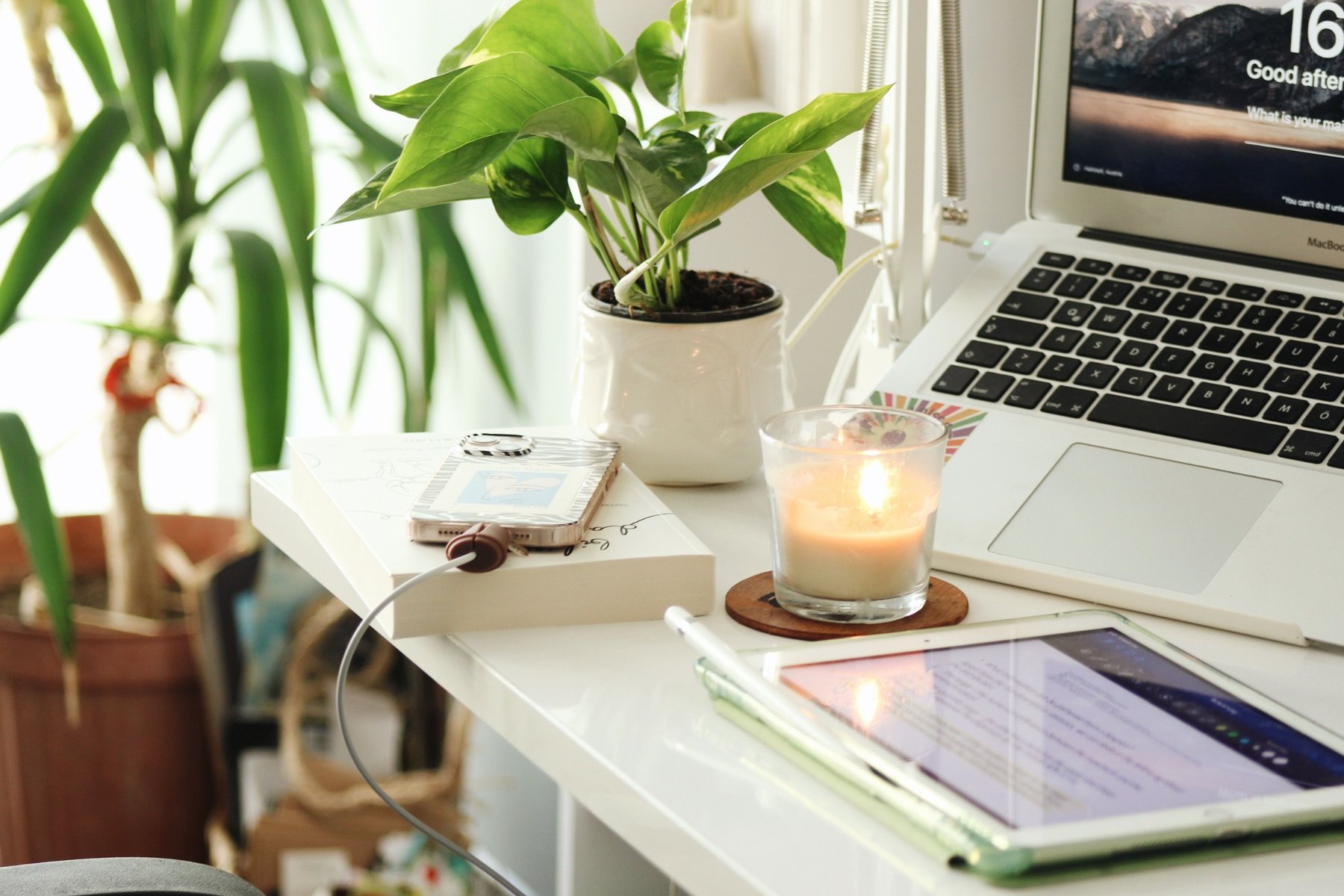 A bright home workspace with a laptop, tablet, phone, candle, and green plants.